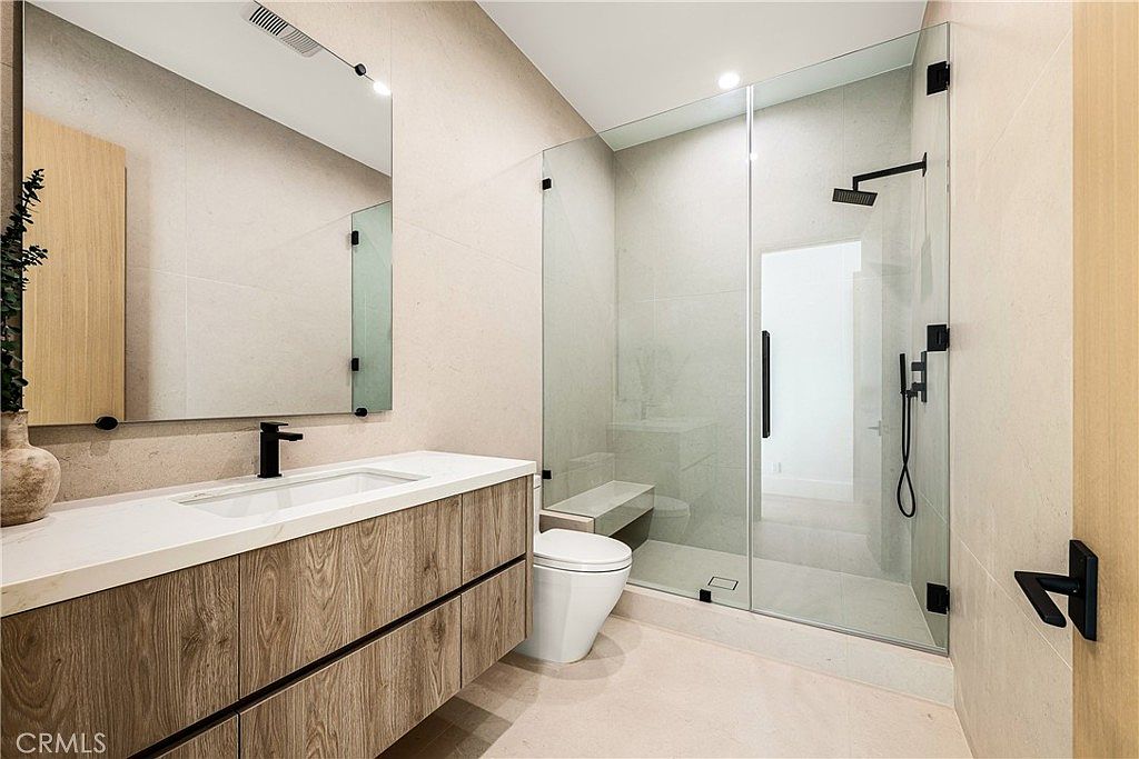This modern, minimalist bathroom features a floating wood-grain vanity with a sleek white countertop and a matte black faucet. A glass-enclosed walk-in shower with a built-in bench and rainfall showerhead adds a luxurious, spa-like feel to the space. The room is finished with large-format neutral tiles, creating a clean, sophisticated, and contemporary aesthetic.