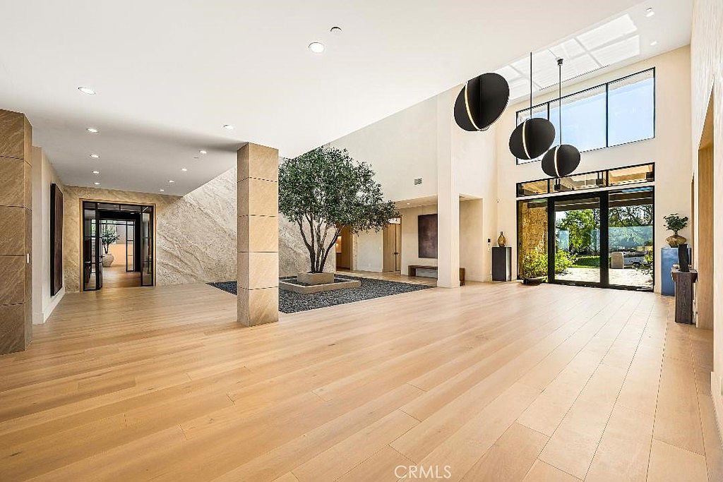 This expansive, high-ceilinged entryway features light-toned hardwood flooring and a striking indoor courtyard with a central tree set in a gravel bed. The space is illuminated by dramatic, oversized spherical pendant lights and large windows that provide a seamless connection to the outdoors. The minimalist design, combined with natural stone accents and clean lines, creates a sophisticated and airy atmosphere.