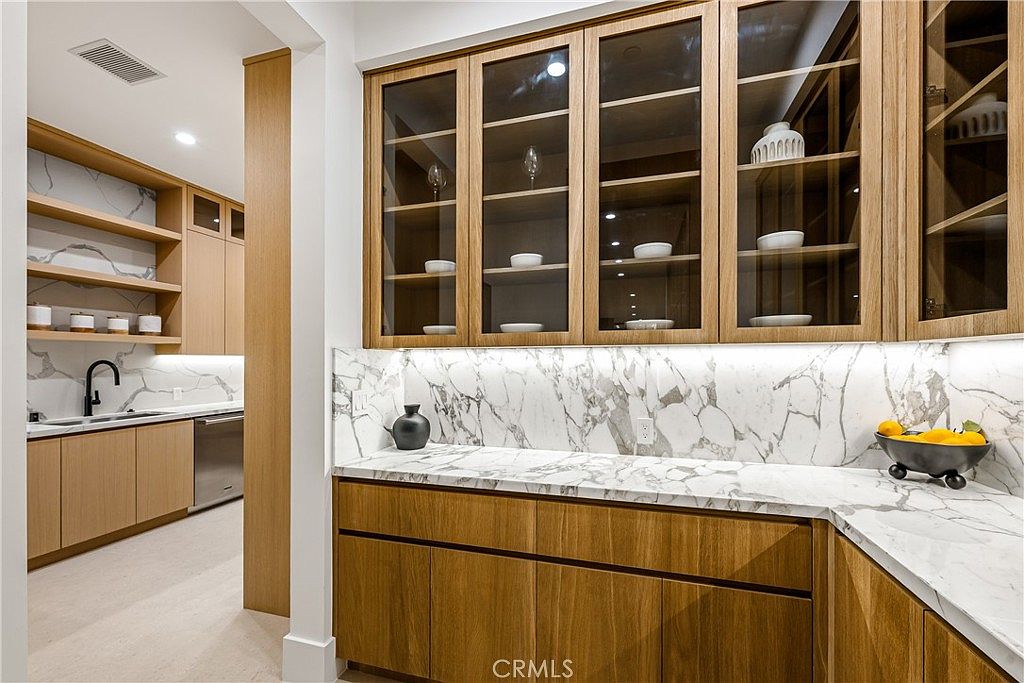 This elegant butler's pantry or secondary kitchen area features warm wood cabinetry paired with striking white marble countertops and a matching backsplash. The space includes glass-front upper cabinets for display, integrated under-cabinet lighting, and a sleek, modern aesthetic. The perspective captures the transition between the pantry area and the main kitchen, highlighting the high-end finishes and clean, organized design.