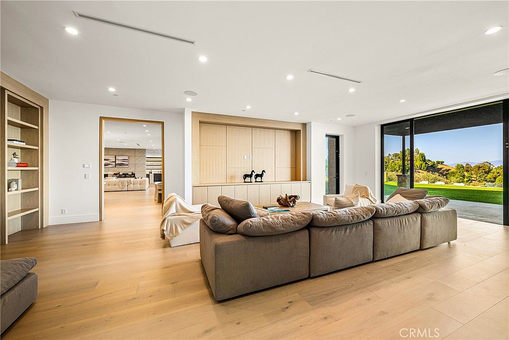 This spacious, modern living room features light hardwood flooring, a large sectional sofa, and a built-in media wall with light wood cabinetry. The room is bathed in natural light from expansive sliding glass doors that open to a scenic outdoor view, creating a seamless indoor-outdoor living experience. The minimalist design and neutral color palette provide an airy, sophisticated atmosphere perfect for relaxation or entertaining.