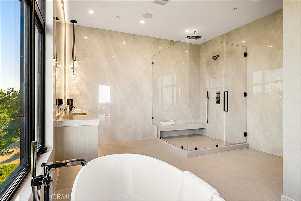 This luxurious primary bathroom features a sleek, modern design with large-format light-toned stone tiles throughout. The space includes a spacious walk-in glass shower with a built-in bench, a contemporary freestanding soaking tub, and a minimalist vanity area illuminated by elegant pendant lighting. Large windows provide ample natural light and a serene view of the surrounding greenery, creating a spa-like atmosphere.