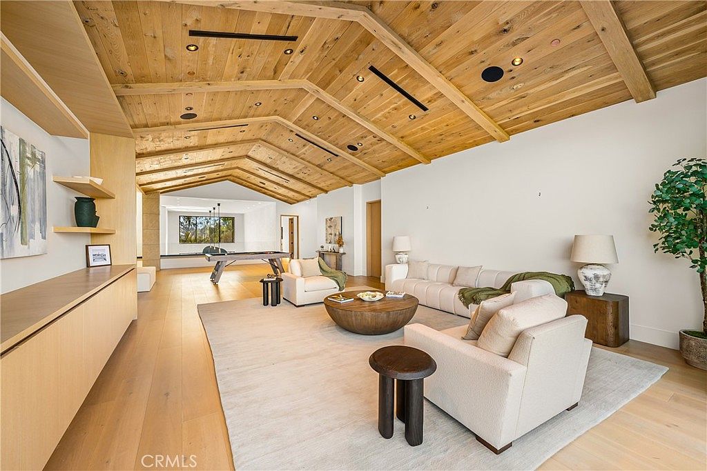 This expansive, open-concept living room features a striking vaulted wood-paneled ceiling with exposed beams and a neutral, modern aesthetic. The space is anchored by a large area rug, a plush white sofa, and matching armchairs, with a circular wooden coffee table serving as a central focal point. In the background, a secondary living area includes a pool table, creating a seamless flow between relaxation and entertainment zones.