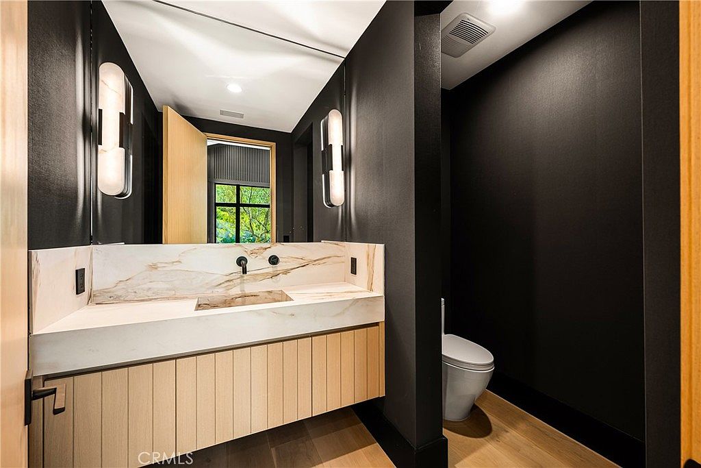 This modern powder room features a striking contrast between dark, matte-black walls and a light wood vanity with a marble countertop. The space is illuminated by two sleek, vertical wall sconces flanking a large mirror, creating a sophisticated and moody atmosphere. A minimalist wall-mounted faucet and a clean-lined toilet complete the contemporary aesthetic.
