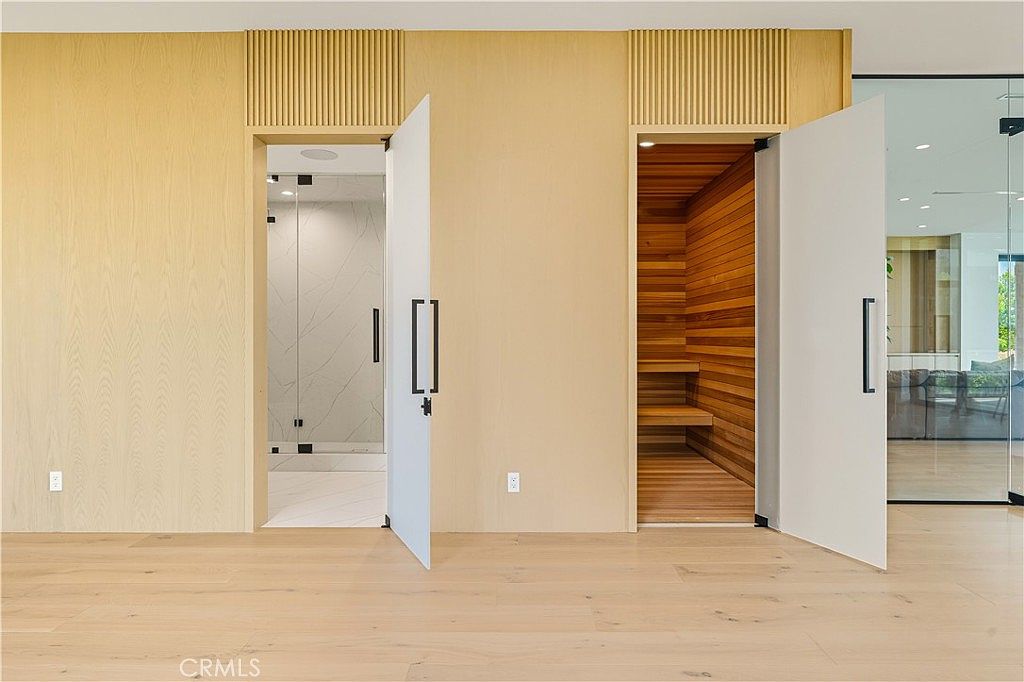 This interior view showcases a minimalist, high-end bathroom area featuring light wood-paneled walls and matching flooring. Two white, floor-to-ceiling doors open to reveal a marble-tiled shower on the left and a warm, cedar-lined sauna on the right. The clean lines and natural materials create a serene, spa-like atmosphere.