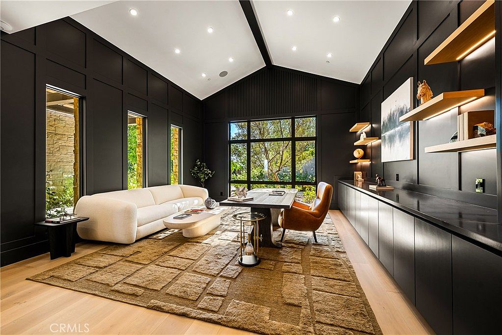 This sophisticated home office features dramatic black paneled walls, a vaulted ceiling, and a large, textured area rug that anchors the space. A sleek desk is paired with a tan leather chair, while a plush white sofa provides a comfortable seating area, all illuminated by recessed lighting and natural light from large windows. The room exudes a modern, high-end aesthetic with floating shelves and built-in cabinetry that offer both style and functionality.
