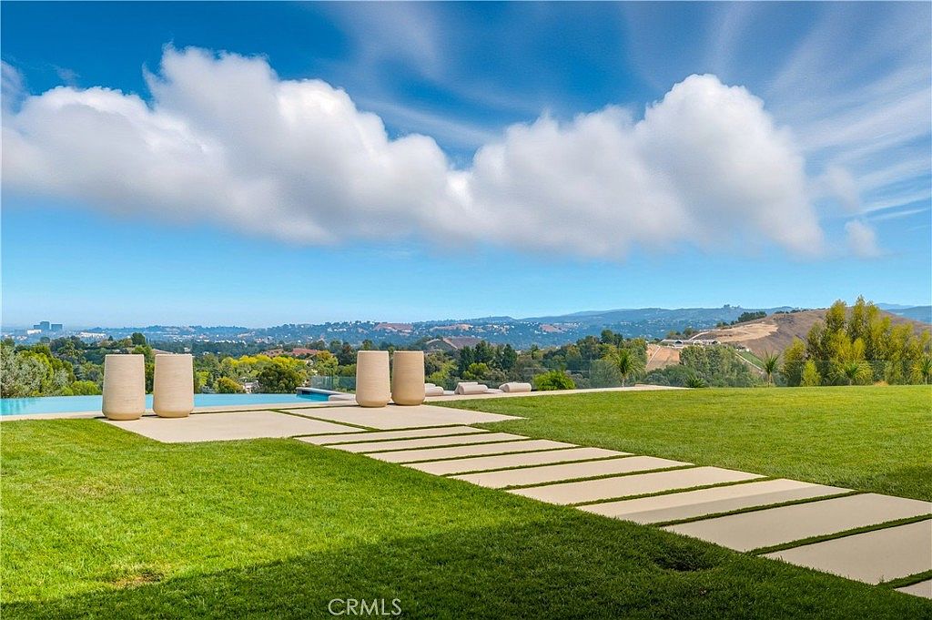 This expansive outdoor space features a sleek infinity-edge pool that seamlessly blends into the rolling hills and distant mountain landscape. A modern stone paver walkway cuts through a lush, manicured lawn, leading toward contemporary cylindrical planters and a serene, open-air setting. The perspective is low and wide, emphasizing the vastness of the property and the tranquil, high-end resort-like atmosphere.