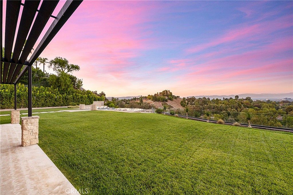 This expansive backyard features a meticulously manicured lawn that stretches toward a stunning panoramic view of rolling hills and a distant cityscape under a vibrant, sunset-hued sky. A modern pergola structure with stone-clad pillars frames the left side of the frame, while a sleek infinity-edge pool sits in the background, seamlessly blending with the horizon. The scene captures a serene, high-end outdoor living space designed for relaxation and taking in the breathtaking natural scenery.