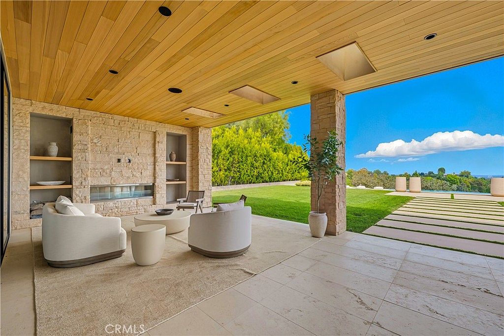 This luxurious covered patio features a seamless indoor-outdoor design with a natural stone fireplace, light-toned tile flooring, and a warm wood-paneled ceiling. The space is furnished with comfortable, modern lounge seating and opens directly onto a manicured lawn and a sparkling pool area, offering a serene view of the surrounding landscape. The overall aesthetic is sophisticated, airy, and perfect for high-end outdoor entertaining.