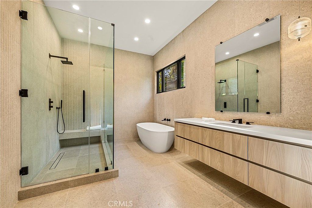 This luxurious primary bathroom features a sleek, modern design with warm, textured stone-look wall tiles and a floating light wood vanity. The space includes a spacious walk-in glass shower, a minimalist freestanding soaking tub, and a large wall-mounted mirror, creating a serene and high-end spa-like atmosphere. The perspective is a wide-angle shot that captures the clean lines and sophisticated material palette of the room.