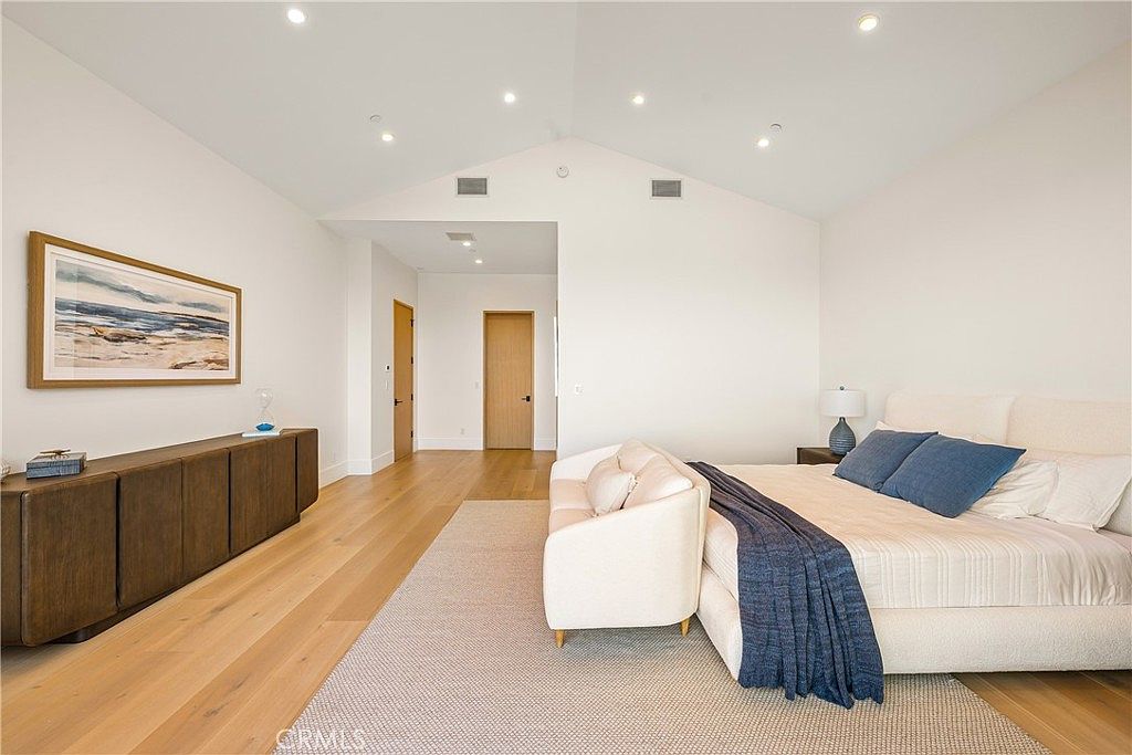 This spacious primary bedroom features a high, vaulted ceiling and light-toned hardwood flooring, creating an airy and serene atmosphere. A large, plush bed with blue accent pillows sits at the center, complemented by a cream-colored sofa at its foot and a long, dark wood console table along the wall. The room is minimalist and modern, accented by a framed coastal landscape painting and recessed lighting that enhances the clean, bright aesthetic.