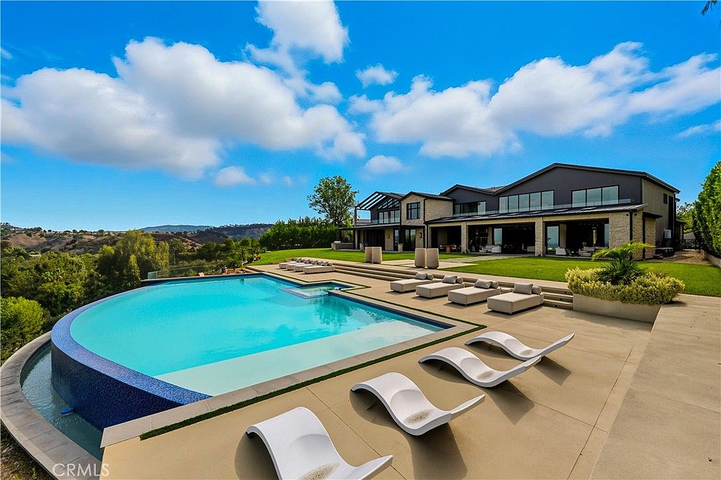 This stunning luxury estate features a large, curved infinity-edge pool that overlooks a vast, rolling landscape under a bright blue sky. The expansive concrete patio is furnished with multiple modern lounge chairs, creating a resort-like atmosphere perfect for outdoor entertaining. The multi-story home showcases contemporary architecture with clean lines, large windows, and a seamless transition between the indoor living spaces and the manicured lawn.