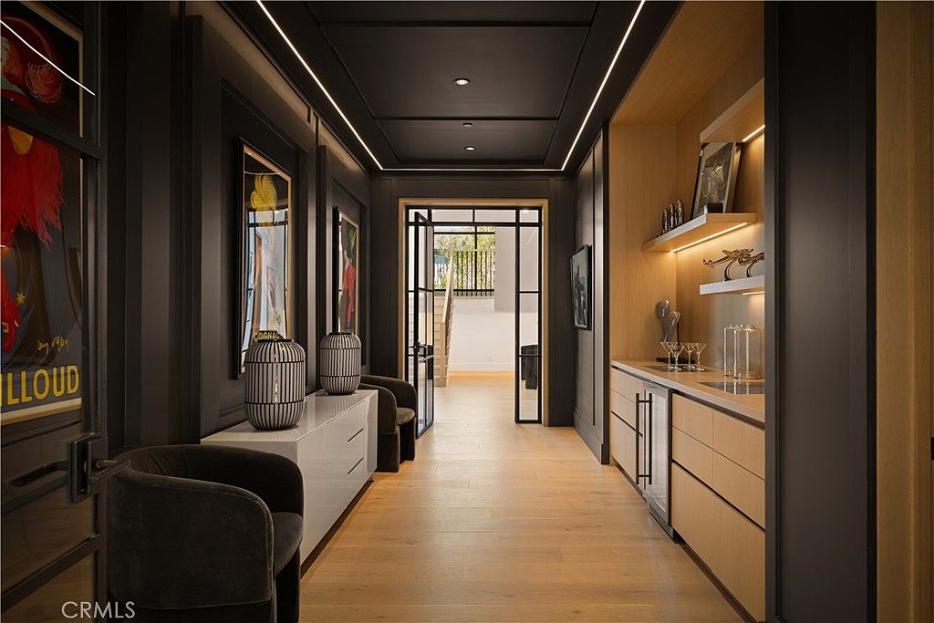 This sophisticated hallway features a dramatic contrast between dark, moody charcoal walls and warm, light-toned wood flooring. The space includes a sleek built-in wet bar with under-cabinet lighting, a modern white console table adorned with decorative lanterns, and framed artwork that adds a touch of personality. The perspective looks down the corridor toward a glass-paned doorway that reveals a glimpse of a staircase, creating a sense of depth and architectural elegance.