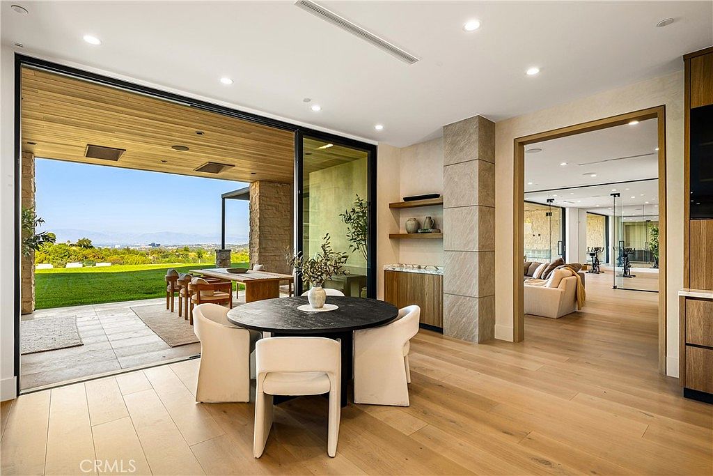 This elegant dining area features a modern circular table with four upholstered chairs, seamlessly transitioning to an expansive outdoor patio through large sliding glass doors. The space is characterized by light hardwood flooring, recessed lighting, and a neutral color palette that creates a bright, airy atmosphere. Beyond the dining area, a glimpse into an adjacent living space and home gym highlights the open-concept layout and luxurious, contemporary design of the home.