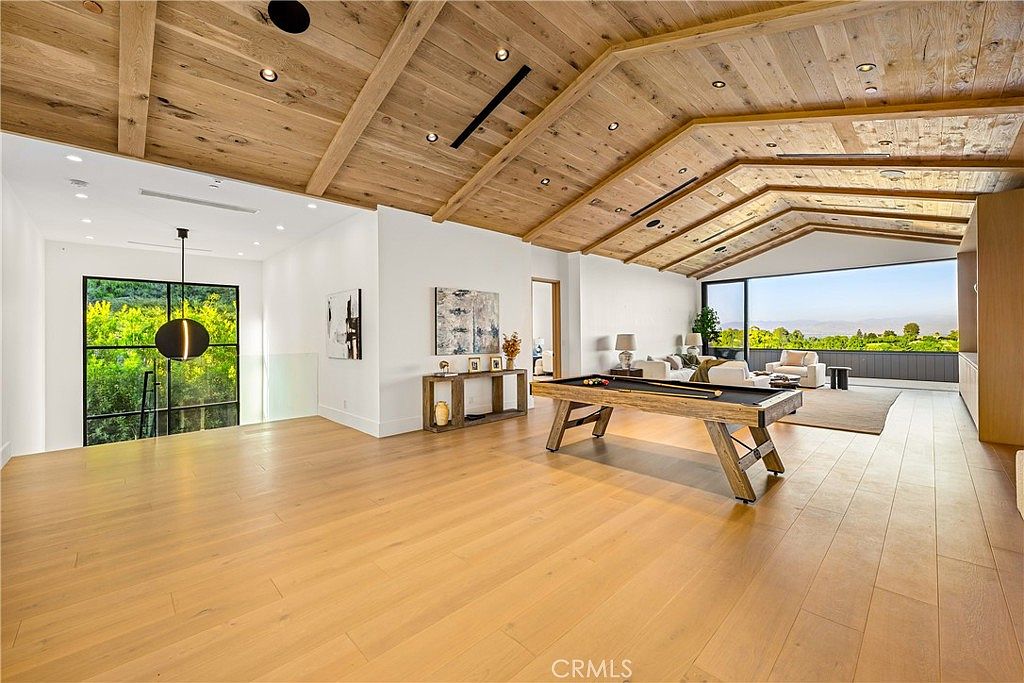 This expansive, open-concept living area features a striking vaulted wood-paneled ceiling and light hardwood flooring, creating a warm and airy atmosphere. The room is anchored by a rustic pool table in the foreground, while the background reveals a comfortable seating area adjacent to a large glass wall that offers panoramic views of the surrounding landscape. The space blends modern architectural design with a relaxed, entertainment-focused layout, perfect for luxury living.