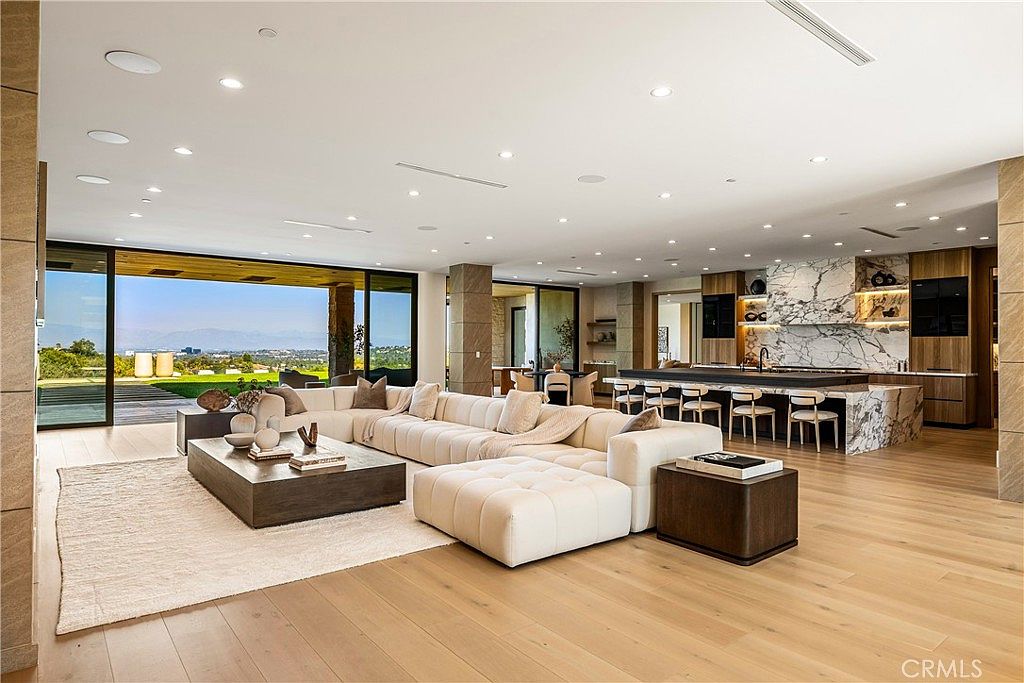 This expansive, open-concept living area features a massive, plush white sectional sofa centered around a low-profile wooden coffee table, creating a luxurious and inviting atmosphere. The space seamlessly transitions into a high-end kitchen with a marble-clad island, while floor-to-ceiling glass walls provide an unobstructed, panoramic view of the outdoor landscape. The design aesthetic is modern and sophisticated, characterized by clean lines, warm wood flooring, and recessed lighting that enhances the airy, bright feel of the room.