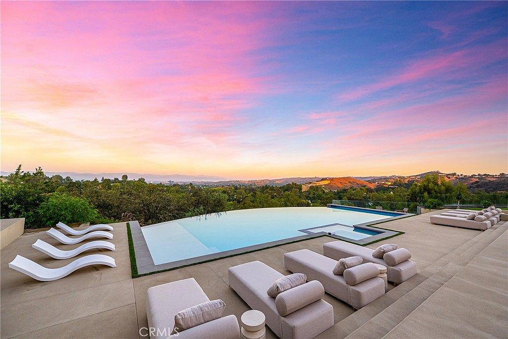 This stunning infinity-edge pool overlooks a vast, rolling landscape under a vibrant, sunset-painted sky. The expansive concrete patio is furnished with multiple modern lounge chairs, creating a luxurious and serene outdoor living space perfect for entertaining. The perspective captures the seamless transition between the pool's edge and the distant horizon, emphasizing the property's high-end, resort-like atmosphere.