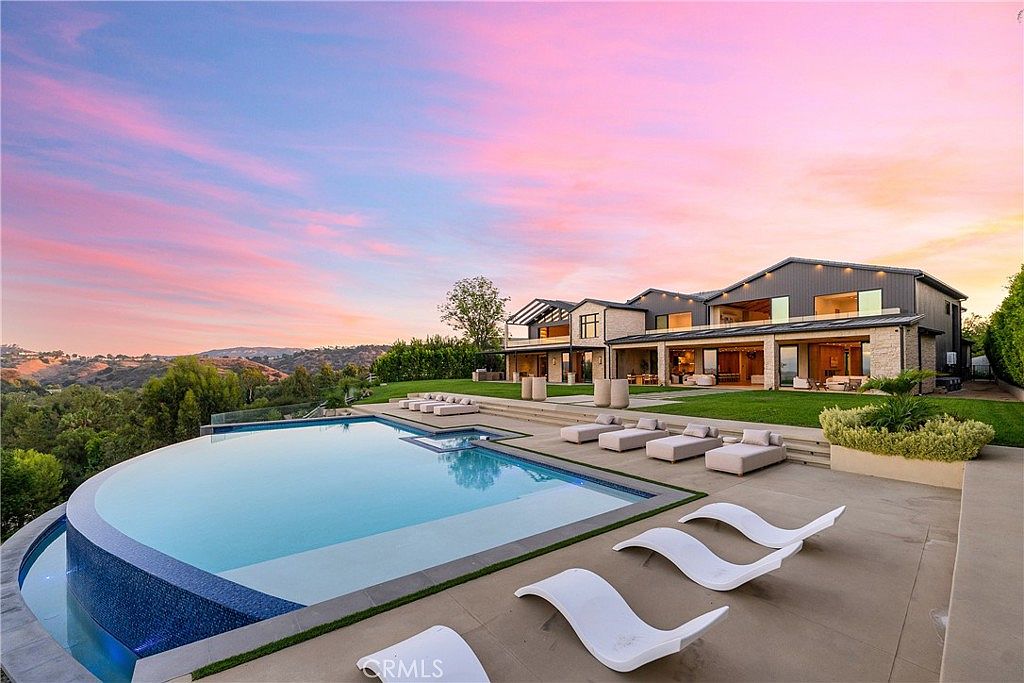 This stunning luxury estate features a sprawling infinity-edge pool that seamlessly blends into the scenic hillside landscape at sunset. The expansive patio area is furnished with modern lounge chairs, creating an ideal space for outdoor entertaining against the backdrop of a vibrant, colorful sky. The architecture of the home is contemporary and sophisticated, with large glass doors that open directly onto the manicured lawn and pool deck.