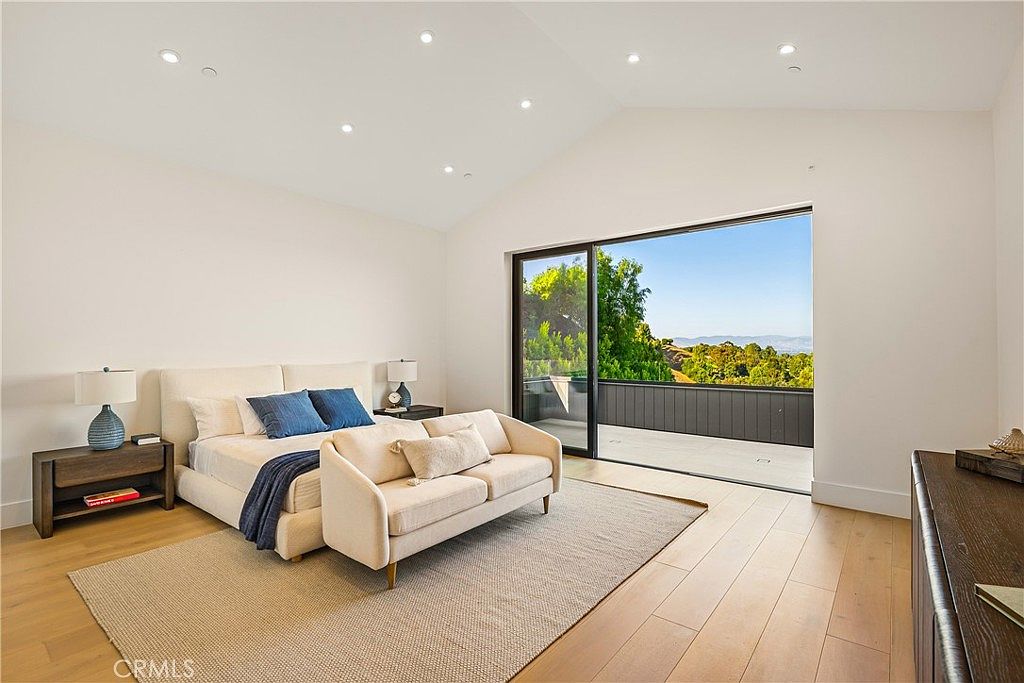 This spacious primary bedroom features a modern, minimalist aesthetic with high vaulted ceilings and light hardwood flooring. A plush bed and a matching sofa are centered on a large area rug, while a wide sliding glass door opens onto a private balcony with scenic hillside views. The room is bathed in natural light, creating an airy and serene atmosphere.