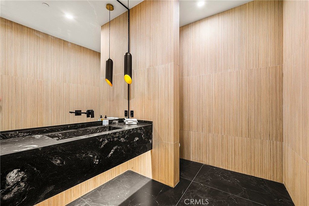 This modern powder room features a striking black marble vanity with an integrated sink and a large, wall-to-wall mirror. The walls are clad in vertical light wood paneling, creating a warm contrast against the dark marble flooring and vanity. Two sleek, minimalist pendant lights hang above the counter, adding a sophisticated and contemporary touch to the space.