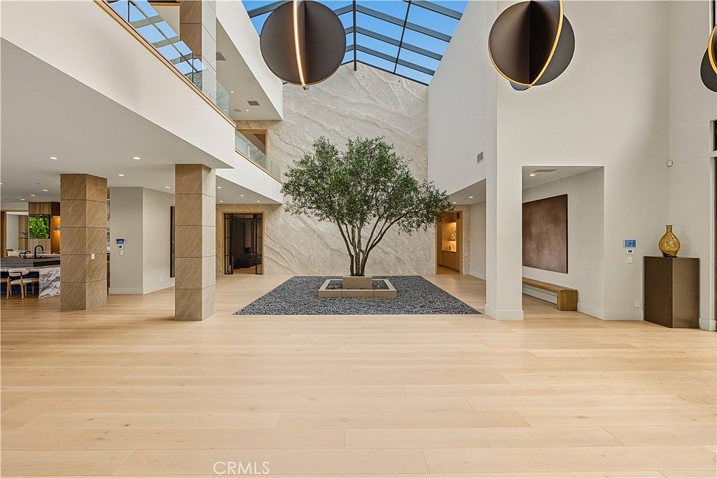 This expansive, high-ceilinged atrium serves as a grand central foyer, featuring light-toned hardwood flooring and a dramatic indoor tree planted in a stone-filled bed. The space is illuminated by a large skylight and modern, oversized spherical pendant lights, creating an airy and sophisticated atmosphere. The architectural design incorporates clean lines, neutral walls, and a visible second-floor balcony, emphasizing a luxurious, open-concept aesthetic.