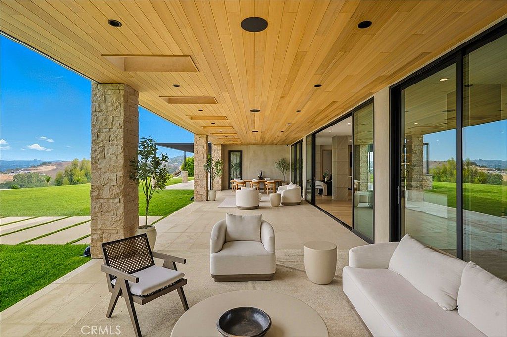 This expansive, covered outdoor patio features a sleek, modern design with a warm wood-paneled ceiling and large stone pillars. The space is furnished with comfortable, neutral-toned lounge seating and a dining area, all seamlessly connected to the interior through floor-to-ceiling glass sliding doors. The patio overlooks a vast, manicured green lawn and rolling hills, creating a serene and luxurious indoor-outdoor living experience.