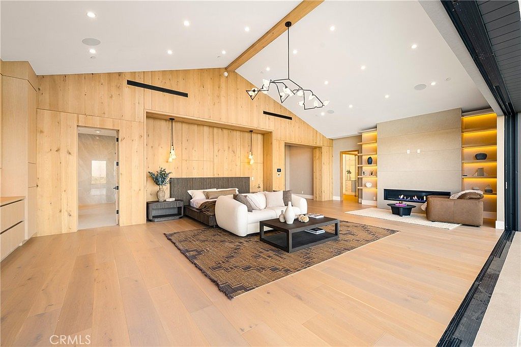 This expansive primary bedroom features a sophisticated, modern aesthetic with warm wood-paneled walls and light-toned hardwood flooring. The room is anchored by a plush white sofa and a central coffee table, creating a cozy seating area in front of a sleek, built-in fireplace and shelving unit. Large, open-concept sliding glass doors seamlessly blend the indoor space with the outdoors, while a contemporary chandelier adds a touch of artistic elegance to the vaulted ceiling.