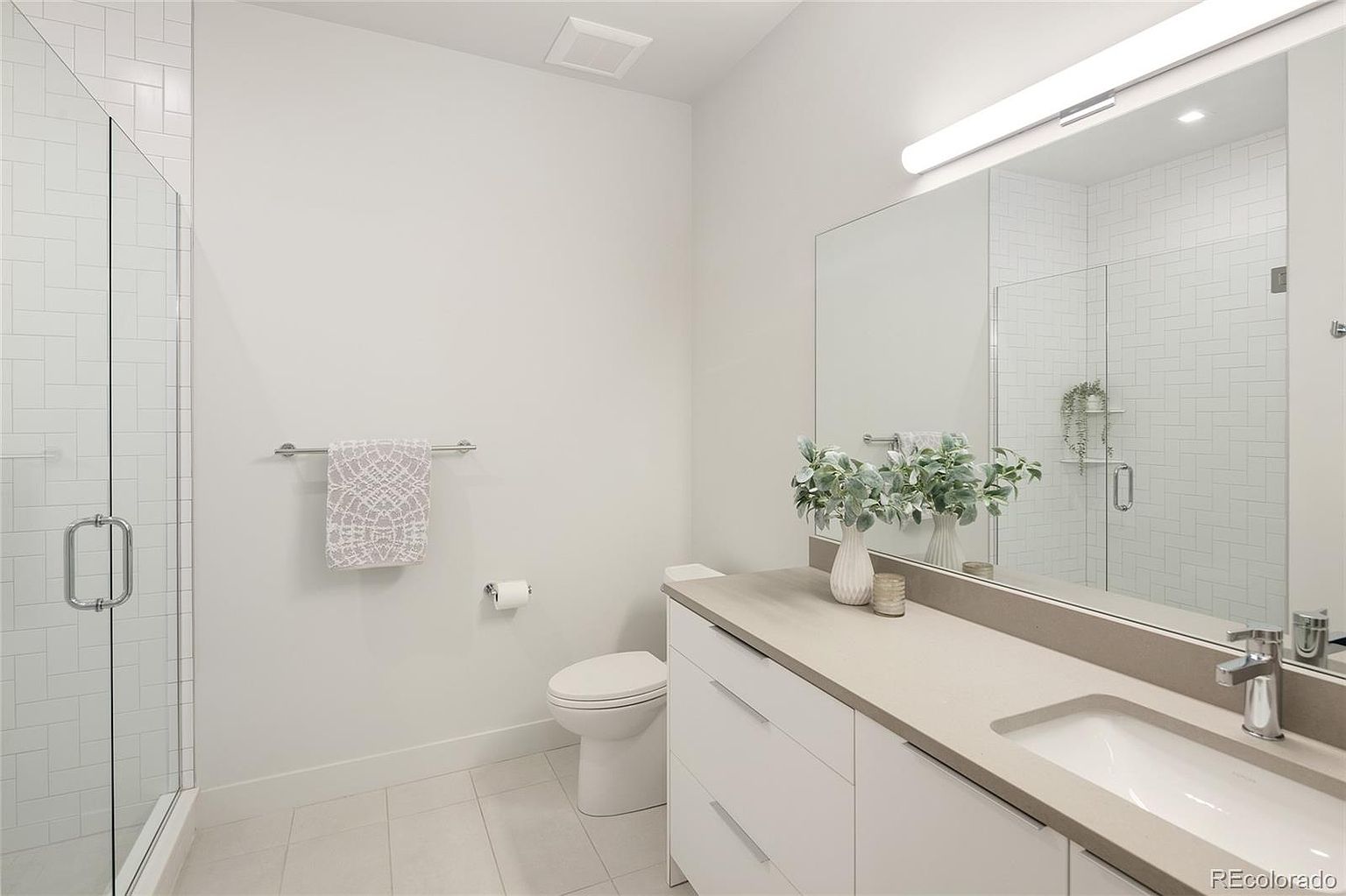 This modern bathroom features a clean, minimalist aesthetic with a white vanity topped by a neutral stone countertop and a large, frameless mirror. To the left, a glass-enclosed shower is finished with white subway tile in a herringbone pattern, while the room is completed by a white toilet and light-colored floor tiles. The perspective is a wide-angle shot from the doorway, capturing the bright, airy atmosphere of the space.