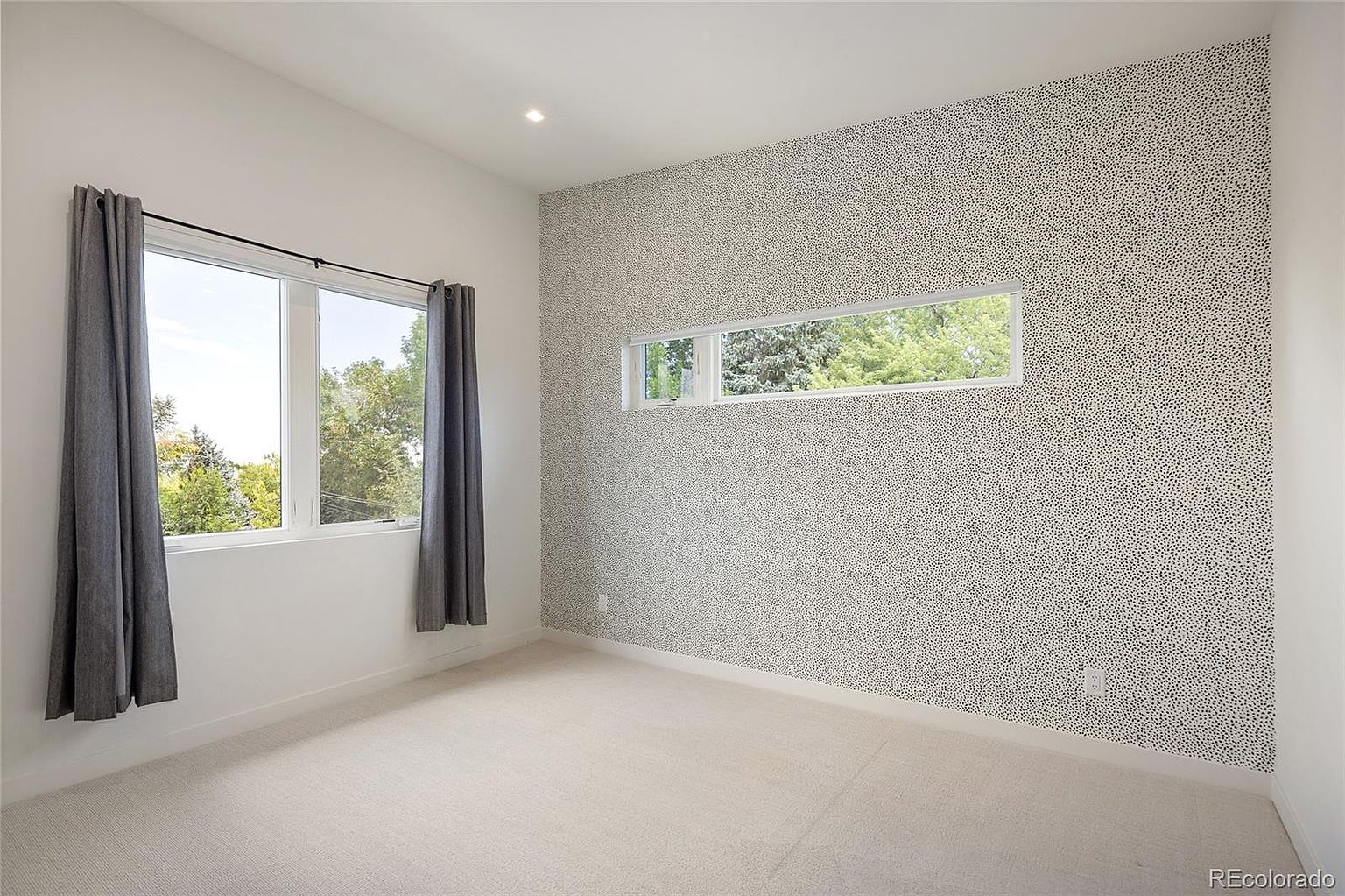 This bright bedroom features a distinctive accent wall with a black-and-white speckled pattern, contrasting with the neutral white walls and light-colored carpeting. The room is illuminated by a large window with grey curtains and a horizontal clerestory window that allows for natural light while maintaining privacy. The space offers a clean, modern aesthetic suitable for a guest room or secondary bedroom.