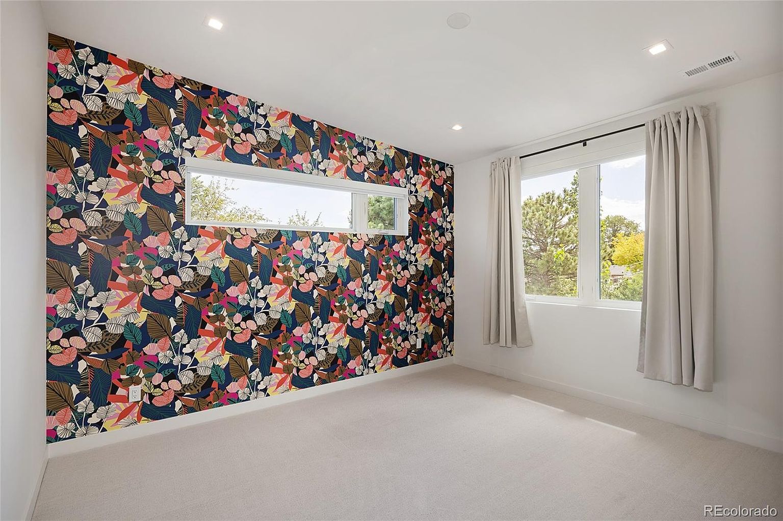 This bright bedroom features a bold, floral-patterned accent wall that serves as a striking focal point against the neutral white walls and carpeting. A long, horizontal window is integrated into the accent wall, while a larger window on the adjacent wall is dressed with simple, light-colored curtains. The room is illuminated by recessed ceiling lights, creating a clean and modern atmosphere.