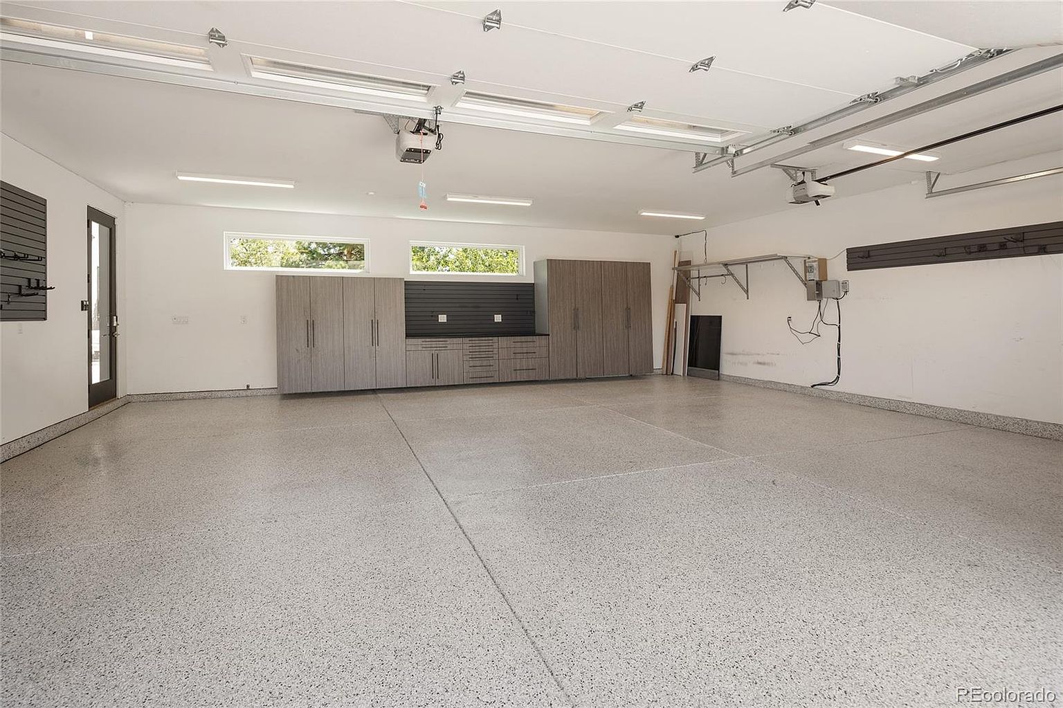 This spacious, well-lit garage features a clean, speckled epoxy floor and bright overhead lighting. A built-in storage system with wood-toned cabinetry and a dark workspace provides organized utility, while two horizontal windows allow natural light to enter the space. The perspective is wide-angle, showcasing the expansive, clutter-free area and the functional design of the storage wall.