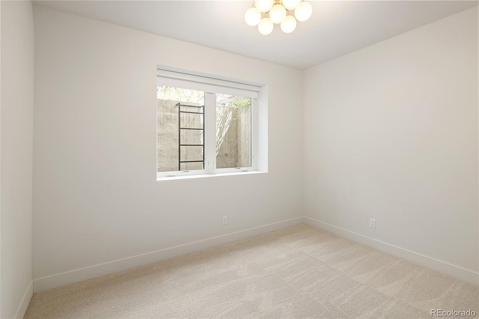 This is a minimalist, neutral-toned bedroom featuring light-colored wall-to-wall carpeting and crisp white walls. A central window with a roller shade provides natural light and a view of an exterior concrete window well with an emergency escape ladder. A modern, multi-globe light fixture hangs from the ceiling, adding a touch of contemporary style to the otherwise simple space.