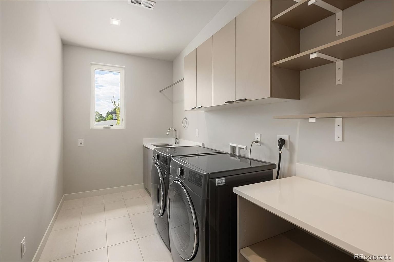 This clean and functional laundry room features a pair of dark-toned front-loading washer and dryer units positioned beneath a white countertop with an integrated sink. The space is equipped with ample storage, including light-colored upper cabinets and open shelving on the side wall. A single window provides natural light, and the room is finished with light-colored tile flooring and neutral wall paint, creating a bright and organized atmosphere.