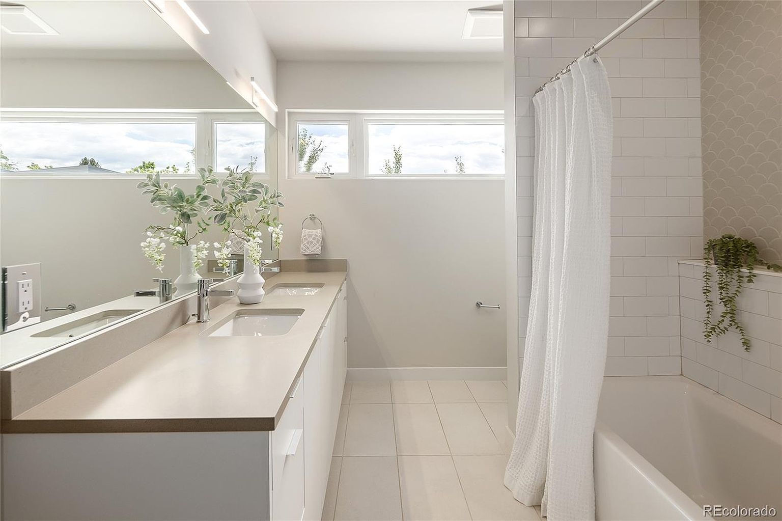 This bright and modern bathroom features a long, dual-sink vanity with a light-toned countertop and a large, expansive mirror that enhances the sense of space. To the right, a bathtub is enclosed by a white shower curtain, complemented by white subway tile walls and a decorative scalloped tile accent wall. The room is illuminated by natural light from high-placed horizontal windows, creating a clean, airy, and contemporary aesthetic.