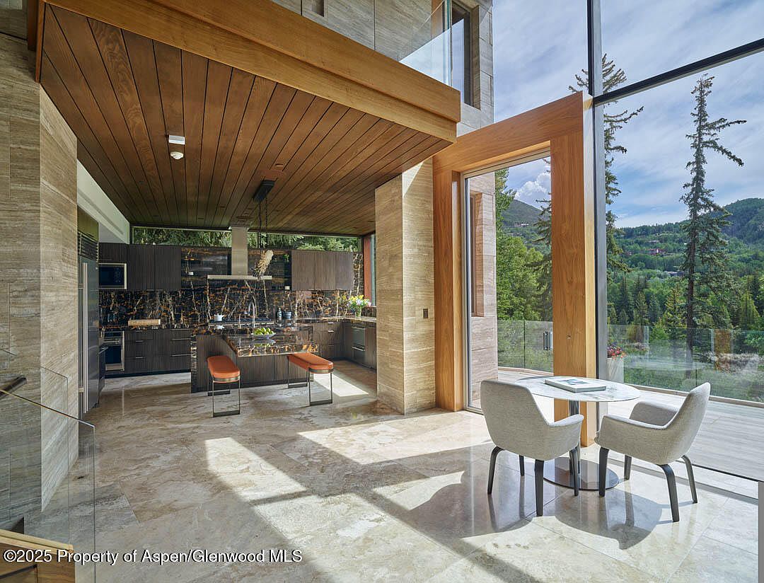 This interior shot showcases a modern kitchen with dark wood cabinetry and marble countertops, complemented by a wooden ceiling and travertine-clad structural elements. A dining area with a round table and two chairs sits adjacent to the kitchen, with floor-to-ceiling windows offering a view of the surrounding landscape. The space is bathed in natural light, highlighting the luxurious finishes and open-concept design.