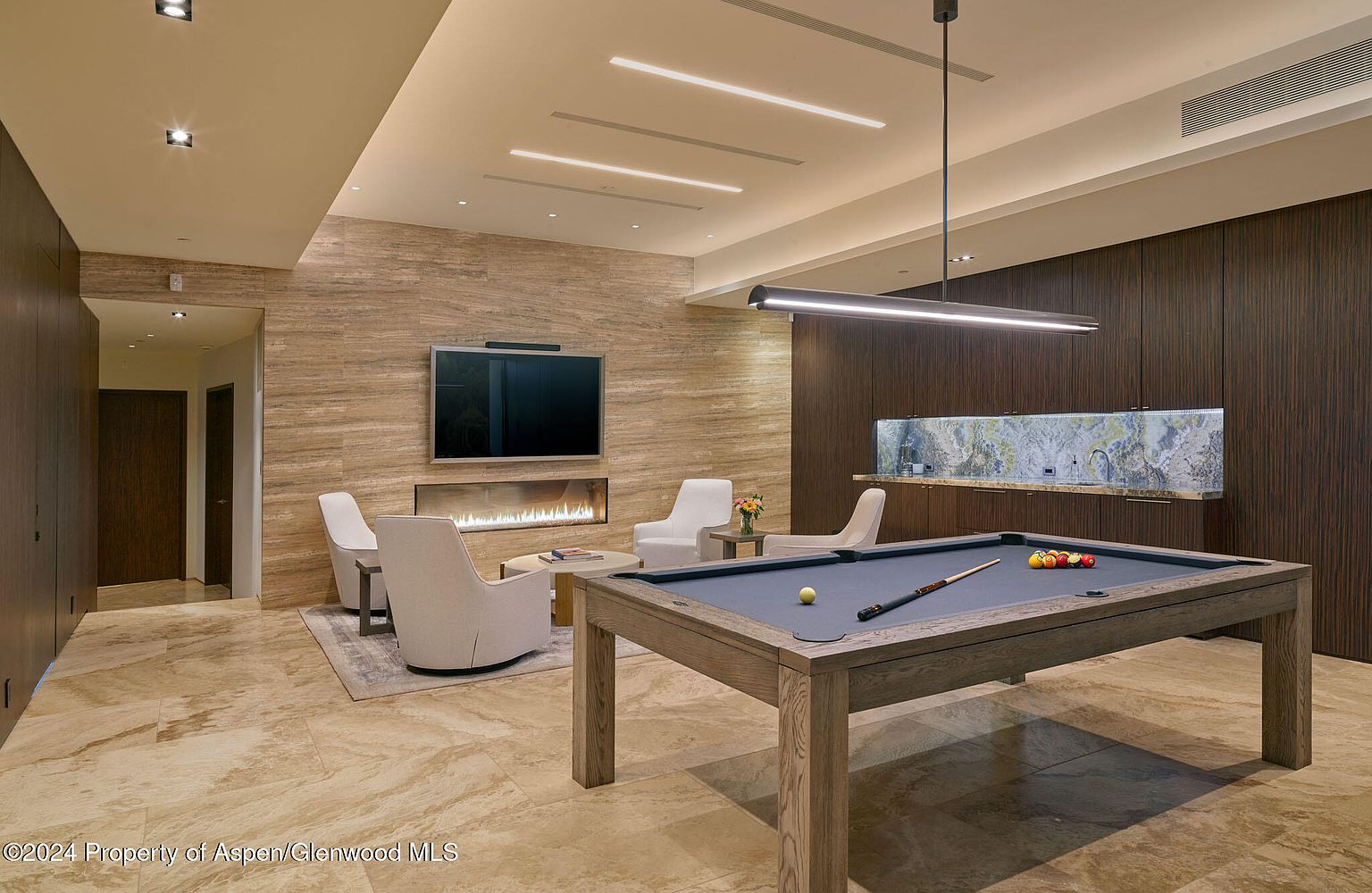This interior shot showcases a luxurious living room featuring a pool table as a central element. The room is adorned with a modern fireplace, a flat-screen TV mounted above it, and comfortable seating arrangements. The design incorporates a blend of textures and materials, including wood paneling and marble flooring, creating a sophisticated and inviting atmosphere.