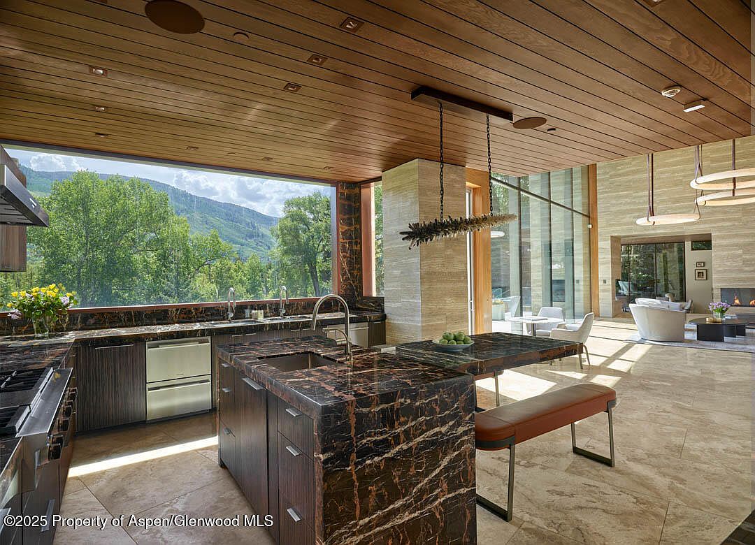This interior shot showcases a modern kitchen with dark, marble-veined countertops and cabinetry, complemented by a wooden plank ceiling. A large window provides a scenic view of the mountains and trees, while a kitchen island with seating extends into an open living space with travertine flooring and contemporary lighting fixtures. The overall impression is one of luxury and seamless indoor-outdoor living.