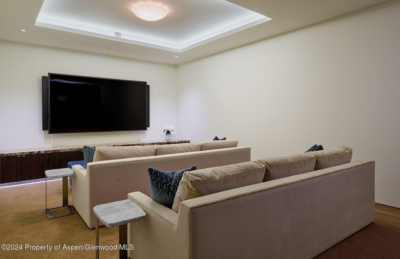 This interior shot showcases a home theater setup with two rows of beige sofas facing a large flat-screen TV. The room features a modern recessed ceiling with soft lighting, and a dark wood cabinet runs along the wall beneath the TV. Small marble-topped side tables are placed next to the sofas, adding a touch of elegance.