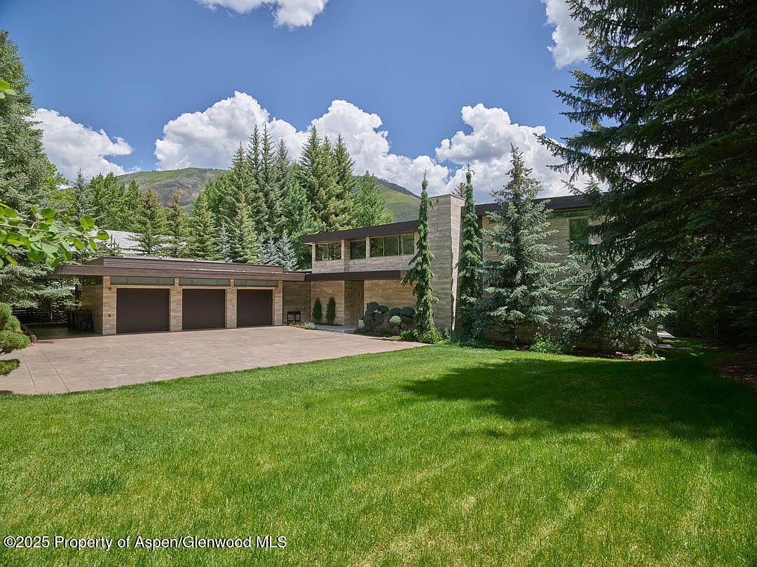 This is a front exterior view of a modern home with a well-manicured lawn. The house features a three-car garage, a stone facade, and large windows. The surrounding landscape includes mature trees and a mountain backdrop, creating a serene and private setting.