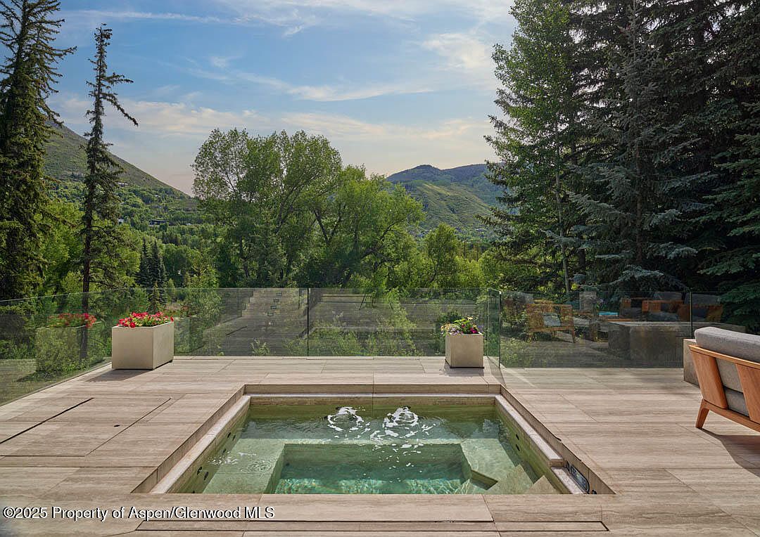 This image showcases a luxurious outdoor patio area with a built-in hot tub as the focal point. The deck is constructed with light-colored wood, and a glass railing provides unobstructed views of the surrounding lush greenery and mountains. The scene exudes relaxation and high-end outdoor living.