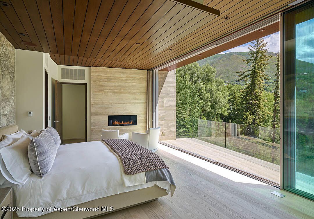 This is a primary bedroom featuring a king-size bed with white linens and decorative pillows. A modern fireplace is set into a stone accent wall, flanked by two white chairs. Large sliding glass doors open to a deck with mountain views, creating a seamless indoor-outdoor living experience.