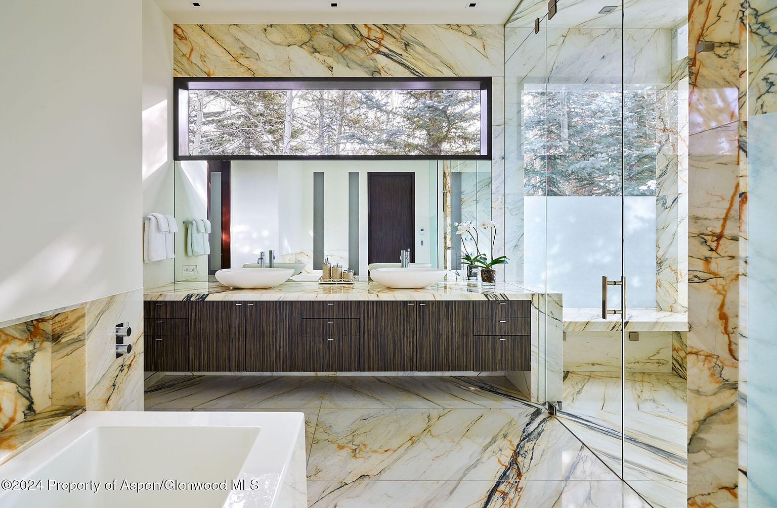 This is a luxurious primary bathroom featuring marble walls and flooring with striking veining. A long, floating vanity with dual vessel sinks sits beneath a large mirror and a horizontal window offering a view of snow-covered trees. A glass-enclosed shower with a built-in bench completes the spa-like atmosphere.