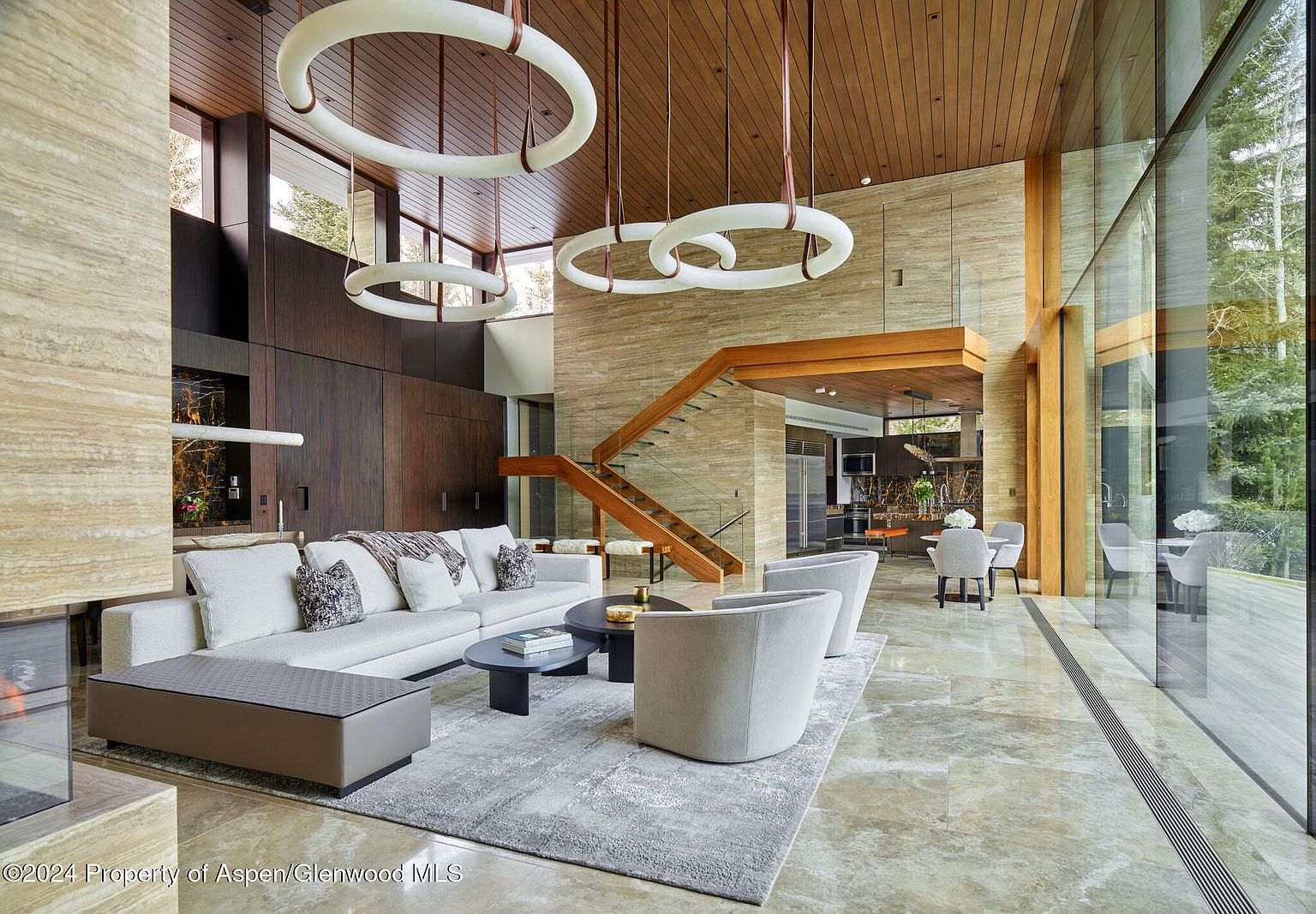 This is a spacious and modern living room featuring a large sectional sofa, two armchairs, and a round coffee table on a gray rug. The room has high ceilings with unique circular light fixtures and a staircase leading to the upper level. Large windows provide ample natural light and views of the outdoors, creating a bright and inviting atmosphere.