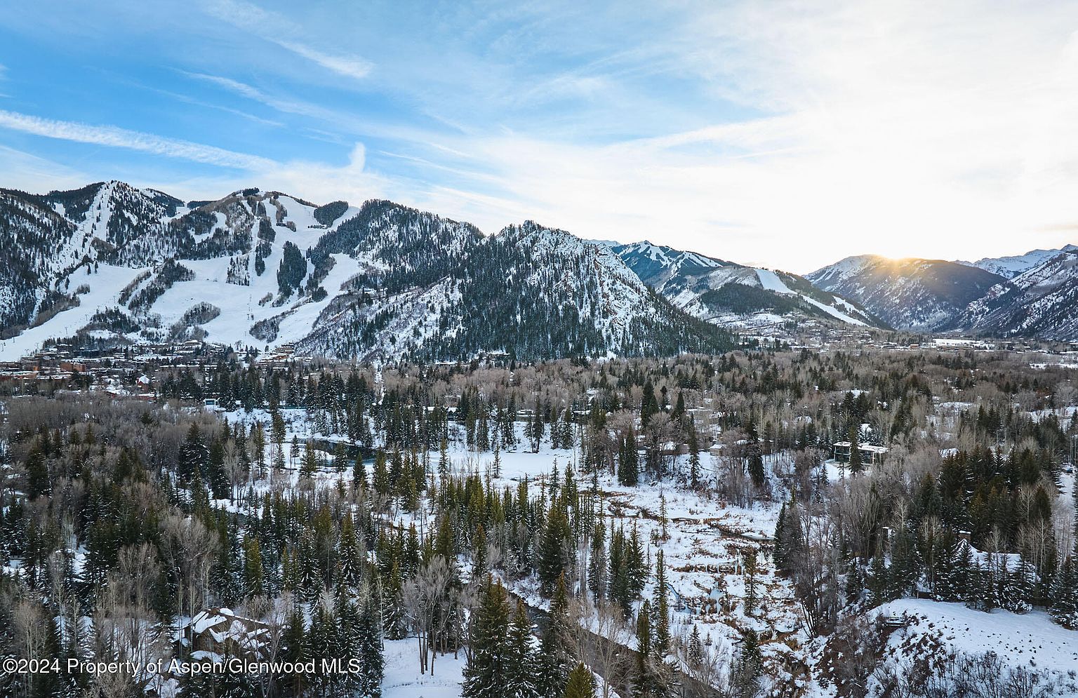 This aerial view showcases a stunning mountain landscape with snow-capped peaks and a lush valley below. The scene features a mix of evergreen forests and snow-covered areas, suggesting a winter or early spring setting. The image provides a sense of grandeur and highlights the natural beauty surrounding the property, emphasizing its desirable location.