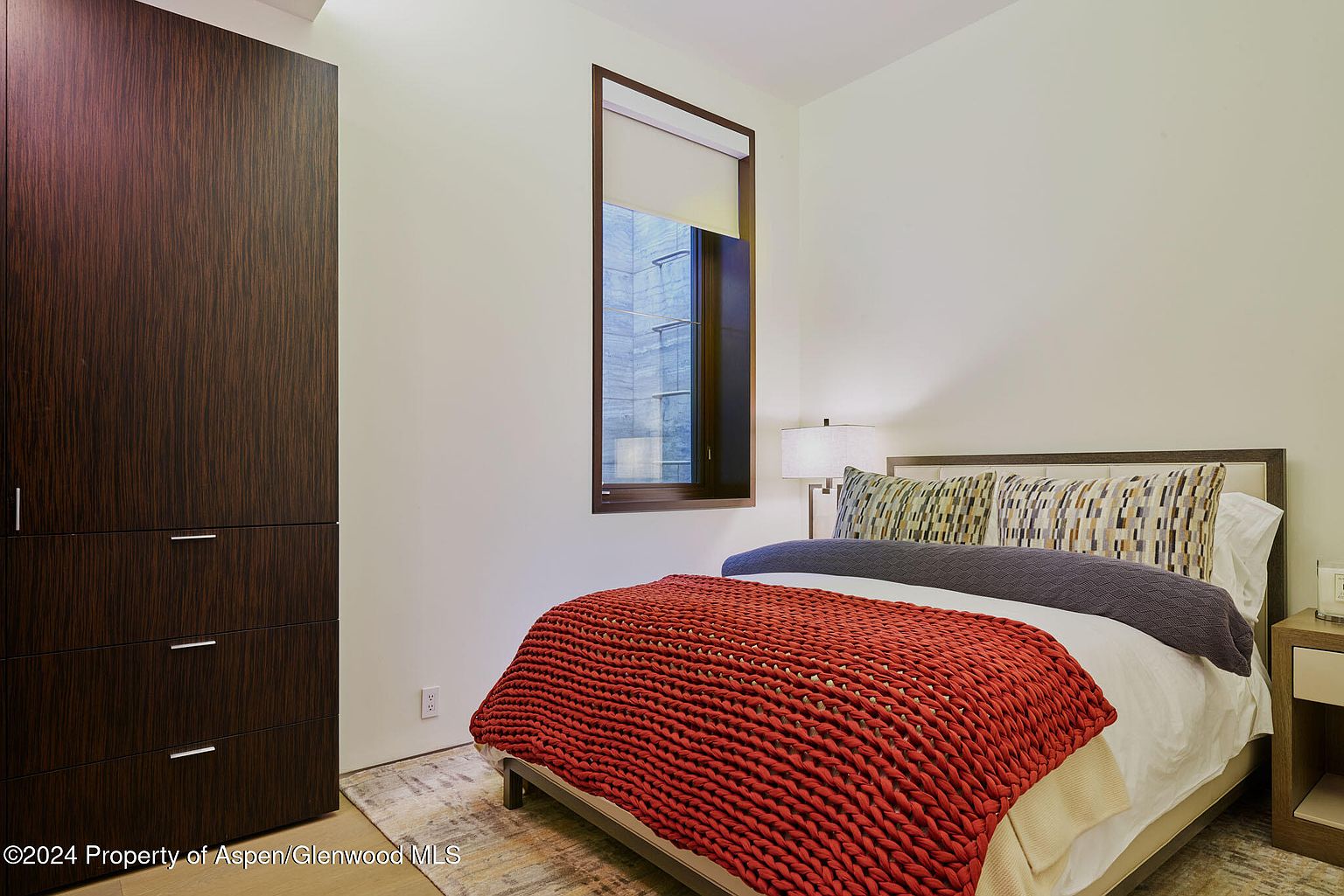 This is a cozy guest bedroom featuring a dark wood wardrobe, a bed with a red knit blanket, and a window with a view of an exterior building. The room has a neutral color palette with white walls and a patterned rug, creating a serene and inviting atmosphere. A bedside table with a lamp adds to the room's functionality and comfort.