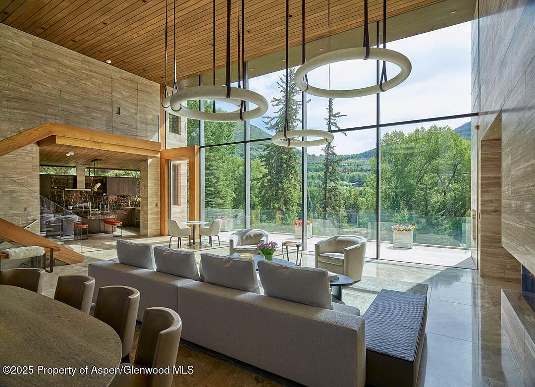 This is a spacious living room with floor-to-ceiling windows offering a view of the surrounding landscape. The room features modern lighting fixtures, a large sofa, and a dining area. The interior design emphasizes natural light and a connection to the outdoors, creating a luxurious and inviting atmosphere.