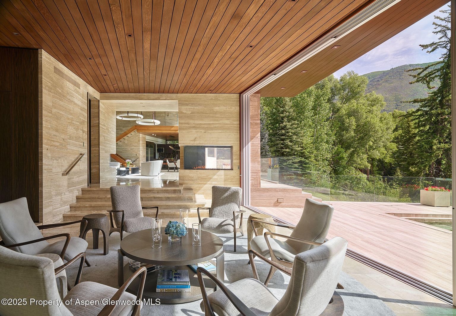 This is an interior shot of a luxurious living room featuring a modern design with natural elements. The room has a seating area with multiple chairs arranged around a round coffee table, a fireplace, and large sliding glass doors that open to an outdoor deck with views of lush greenery. The use of wood and stone textures creates a warm and inviting atmosphere.