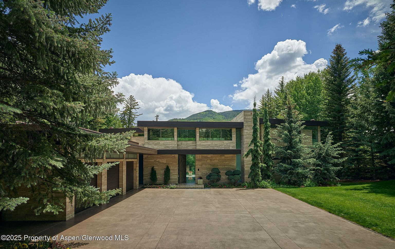 This is a front view of a modern, two-story home with a stone facade and dark-framed windows. The property features a large concrete driveway, manicured landscaping with mature trees, and a glimpse of mountains in the background. The architecture emphasizes clean lines and a seamless integration with the natural surroundings, creating an impression of sophisticated tranquility.