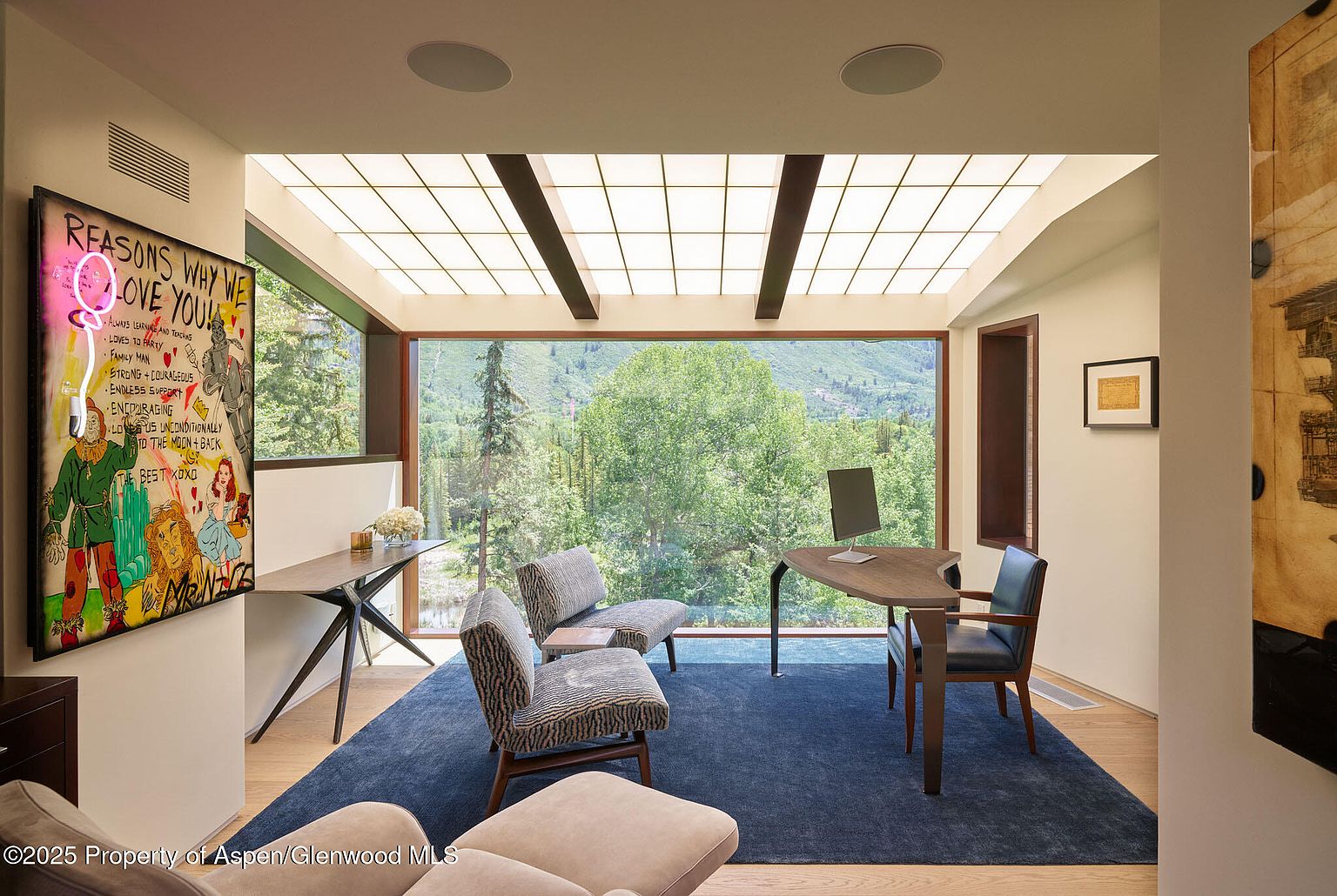 This is a bright and airy office space featuring a large window with a view of lush greenery. The room is furnished with a modern desk, comfortable chairs, and a vibrant piece of art. A blue rug anchors the space, creating a cozy and inviting atmosphere perfect for productivity.