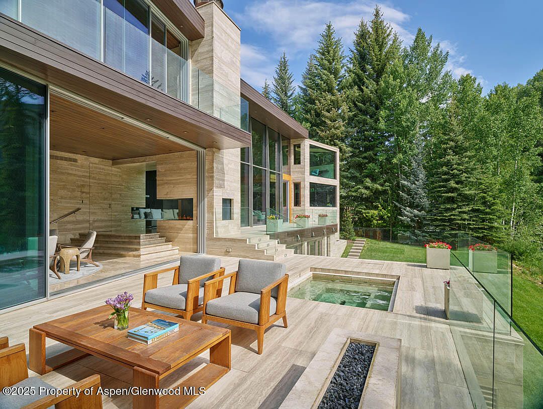 This image showcases a luxurious outdoor patio area of a modern home. The patio features a wooden deck with stylish outdoor furniture, including chairs and a coffee table. A small pool or spa is visible, along with a fire pit and glass railings that offer unobstructed views of the surrounding lush greenery and trees. The architecture is contemporary, with clean lines and a seamless integration of indoor and outdoor spaces.