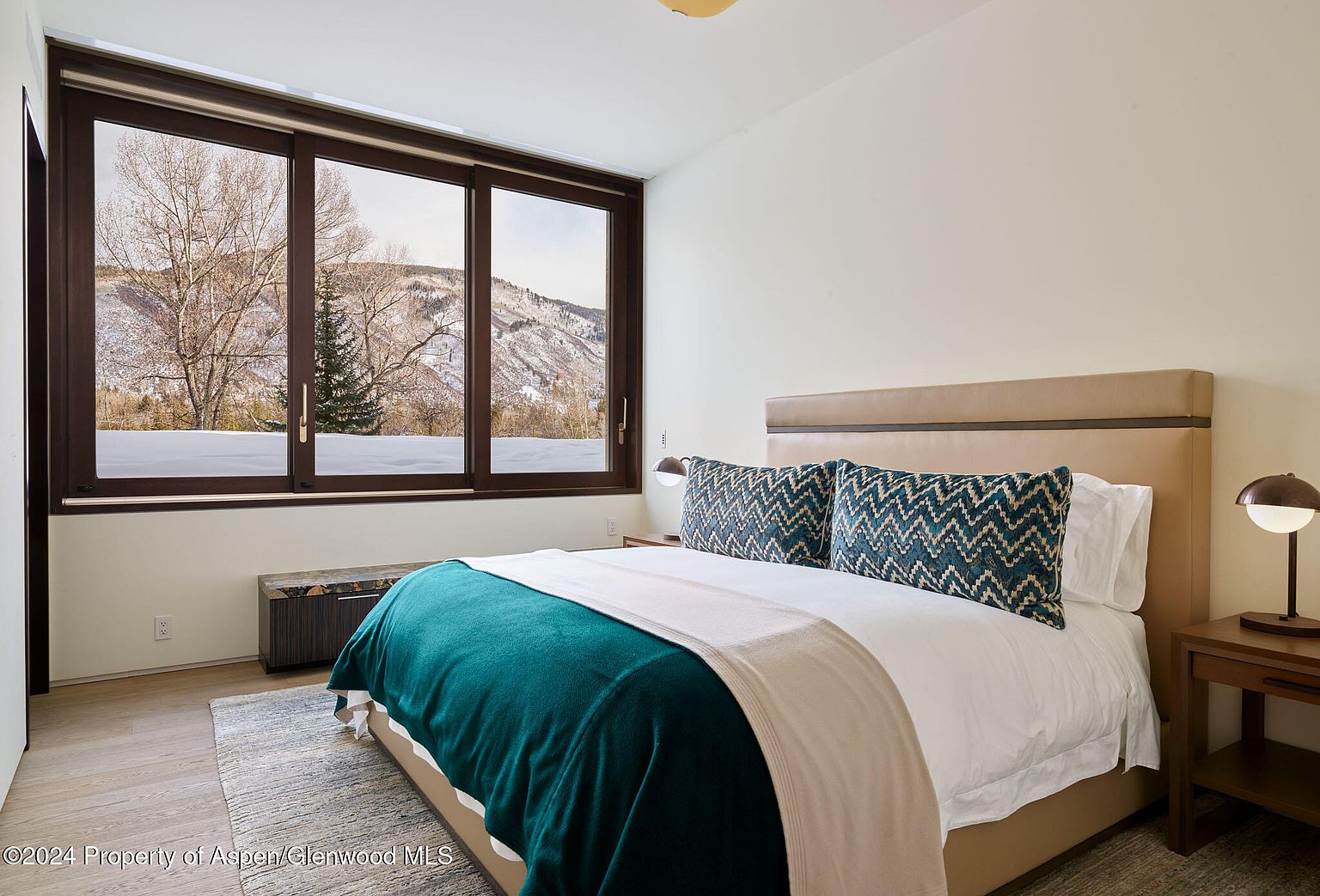 This is a cozy bedroom featuring a large window with a view of snow-covered mountains. The bed is neatly made with a teal blanket, a beige throw, and patterned pillows. A wooden nightstand with a lamp sits beside the bed, adding a warm glow to the room. The overall impression is one of comfort and tranquility.